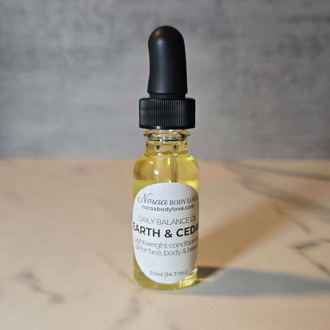 Noraa Body Love Earth & Cedar Daily Balance Oil in a clear glass dropper bottle with black top, featuring a simple label, placed on a neutral background.