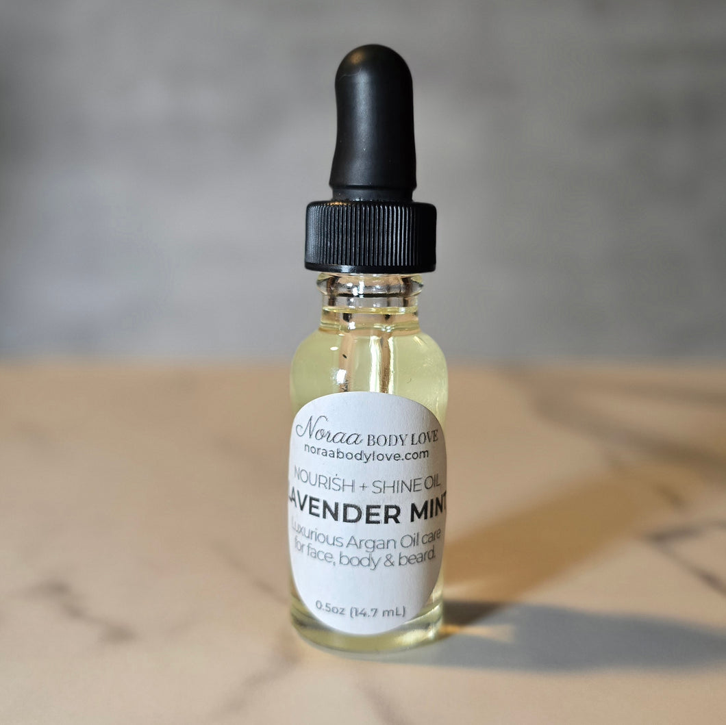Noraa Body Love Lavender Mint Nourish + Shine Oil in a clear bottle with a black dropper, labeled for face, body, and beard. Displayed on a marble surface.
