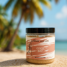 Load image into Gallery viewer, A jar of Noraa Body Love Coco Bay Sugar Soap Scrub sits on soft sand with a blurred backdrop of palm trees and coconuts. The label is centered and easy to read, and the tropical setting highlights the beachy scent theme.
