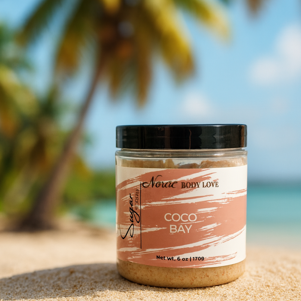 A jar of Noraa Body Love Coco Bay Sugar Soap Scrub sits on soft sand with a blurred backdrop of palm trees and coconuts. The label is centered and easy to read, and the tropical setting highlights the beachy scent theme.