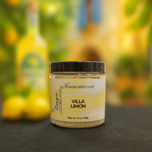 Load image into Gallery viewer, A jar of Noraa Body Love&#39;s Villa Limon Sugar Soap Scrub sits on a black surface with a softly blurred limoncello-inspired background featuring hints of lemons, greenery, and warm yellow tones
